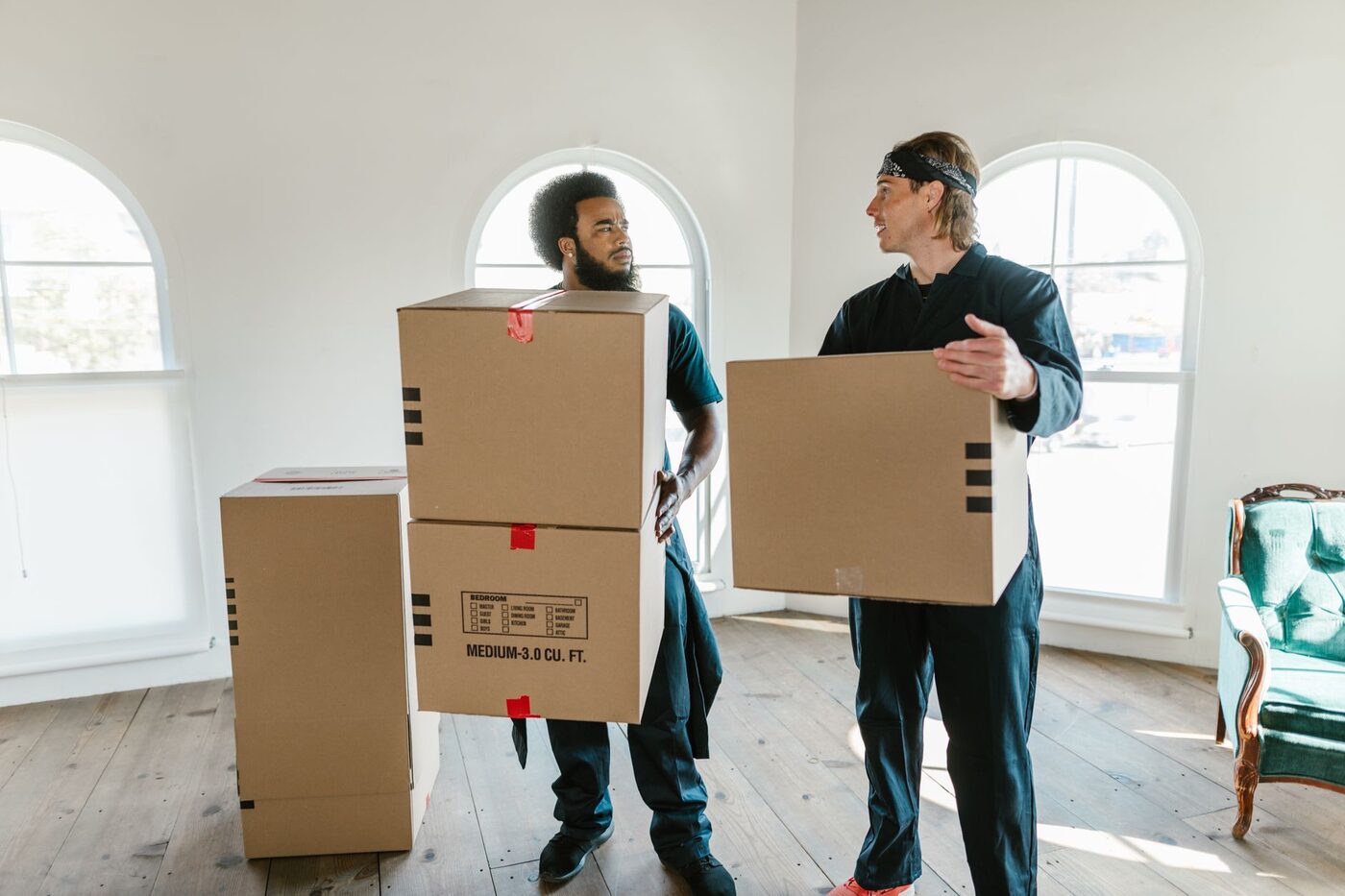 Two movers in uniform carrying cardboard boxes through a bright empty room
