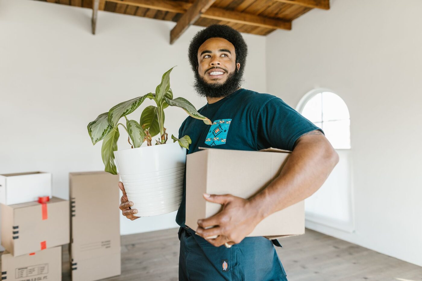 A mover smiling while carrying a box and a potted plant into a bright new home