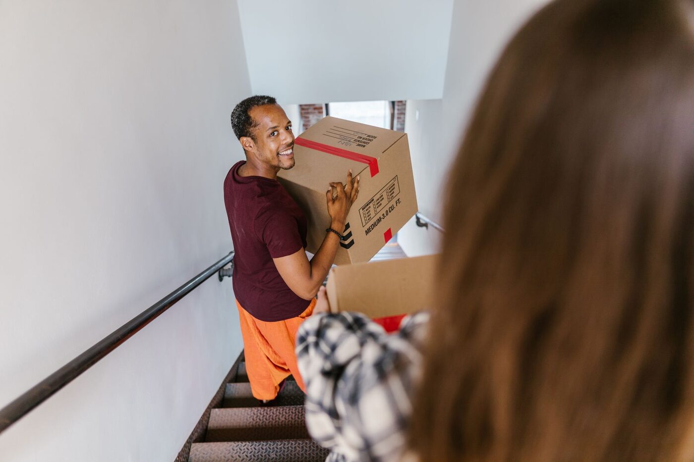A man smiling while carrying a moving box up an indoor staircase